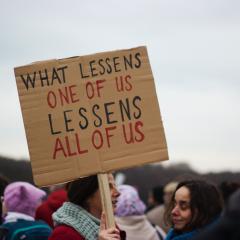 Protesting woman holding up a sign saying "what lessens one of us, lessens all of us" - credit Micheile Henderson