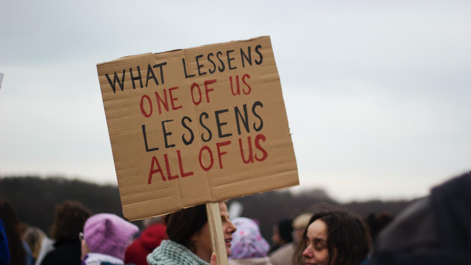 Protesting woman holding up a sign saying "what lessens one of us, lessens all of us" - credit Micheile Henderson
