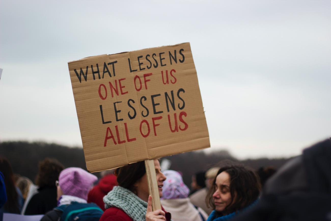 Protesting woman holding up a sign saying "what lessens one of us, lessens all of us" - credit Micheile Henderson